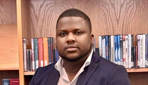 Thélyson Orélien devant une bibliothèque, vêtu d'un blazer marine.