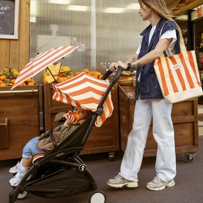 Femme en tenue décontractée (jean, blanc) promenant un enfant en poussette dans un marché.