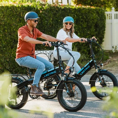 Un couple circulant à bord de vélos électriques Lectric sur une piste pavée bordée de haies vertes.