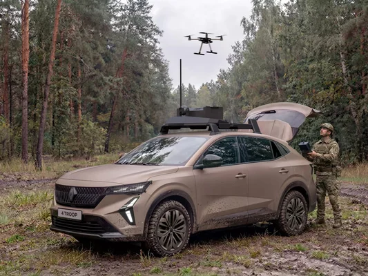 Un soldat en tenue de camouflage utilise une manette pour piloter un drone, aux côtés d'un SUV militaire doté d'antennes, en forêt.