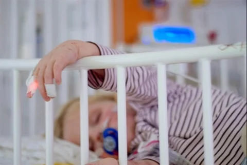 Jeune enfant endormi dans un berceau, avec une main d'adulte tenant un dispositif médical, évoquant les soins pédiatriques.