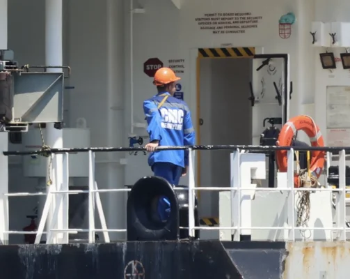 Ouvrier en uniforme bleu et casque orange sur le pont d'un navire, près d'une porte de sécurité.