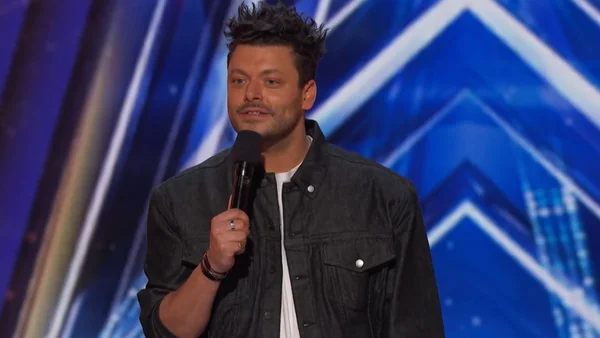 Kev Adams sur scène, micro à la main, devant un décor bleu.
