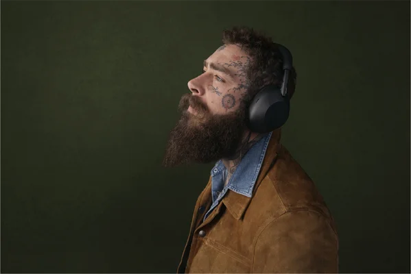 Homme barbu aux tatouages faciaux portant le casque Sony WH-1000XM6, veste en daim et chemise en jean.