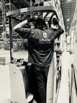 Travailleur conduisant un chariot élévateur dans un entrepôt, arborant le logo UNIFOR sur sa chemise.