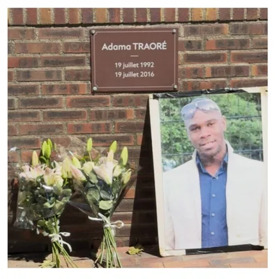 Hommage à Adama Traoré : plaque, fleurs et portrait sur un mur de brique.