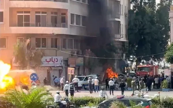 Incendie et fumée noire près de l'hôtel de Macron à Damas, avec des pompiers et des badauds sur place.