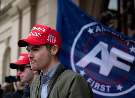 Nick Fuentes arborant une casquette MAGA devant un drapeau America First.