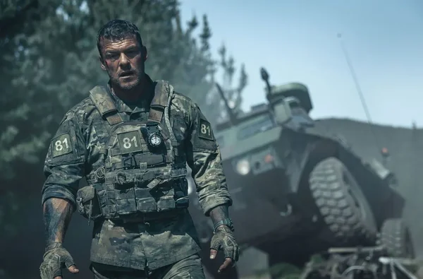 Soldat en tenue de combat dans une forêt avec un véhicule blindé en arrière-plan (War Machine).