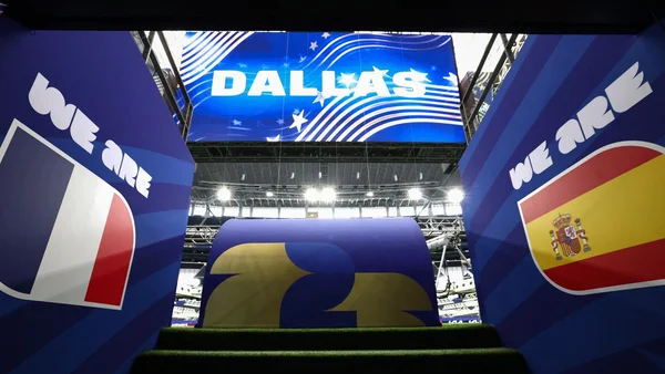 Cette image montre une vue de l'intérieur d'un tunnel de stade donnant sur le terrain, avec de grandes banderoles bleues de chaque côté affichant 'WE ARE' accompagnées des drapeaux français et espagnols. Un grand écran en arrière-plan affiche 'DALLAS' avec des graphiques en étoile, indiquant le lieu de cet événement sportif.