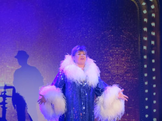 Michèle Bernier dans son spectacle Vive demain! sur scène, en tenue bleue à paillettes et plumes blanches.