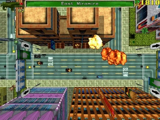 In-game screenshot from Grand Theft Auto (1997), showing top-down 2D gameplay with pixel-art graphics, cars on city streets, explosions, and UI elements like score and location name ('East Miramire').