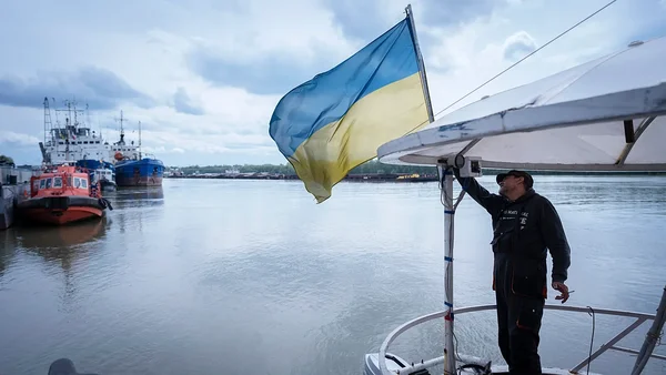 Un marin ukrainien brandissant le drapeau national sur un navire dans un port, sous un ciel couvert.
