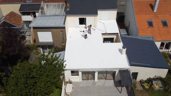 Deux ouvriers peignant un toit plat en blanc dans un quartier résidentiel, une pratique qui séduit particuliers et villes.