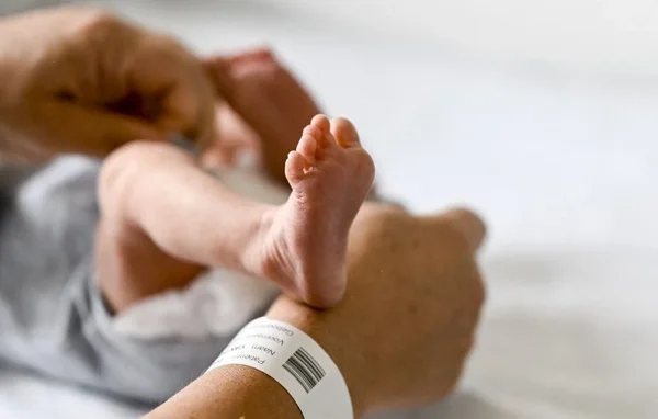 Le pied d'un nouveau-né reposant sur un poignet adulte avec un bracelet d'hôpital, symbole de la naissance en période de canicule.