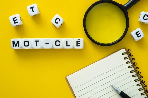 Cubes de lettres formant le mot 'MOT-CLÉ' sur fond jaune, avec une loupe et un carnet.