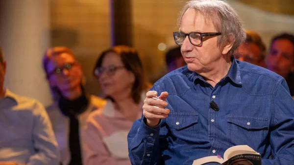 Michel Houellebecq prenant la parole lors d'un débat, un livre ouvert à la main.