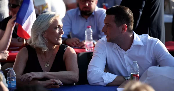 Marine Le Pen et Jordan Bardella en conversation lors d'un événement en extérieur, avec des drapeaux russes visibles.
