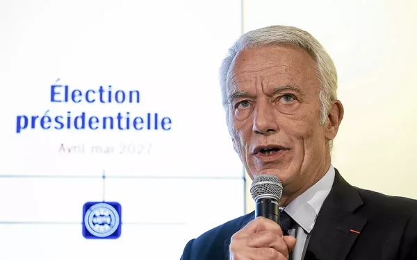 Homme en costume parlant dans un micro devant un fond 'Élection présidentielle'.