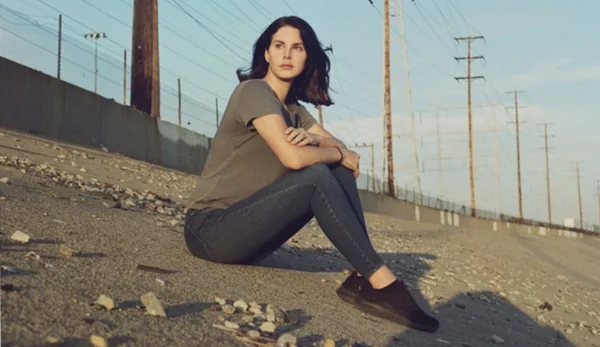 Lana Del Rey assise sur un talus graveleux dans un décor industriel.