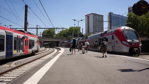 Quai de gare animé par une belle journée, avec des trains et des voyageurs.
