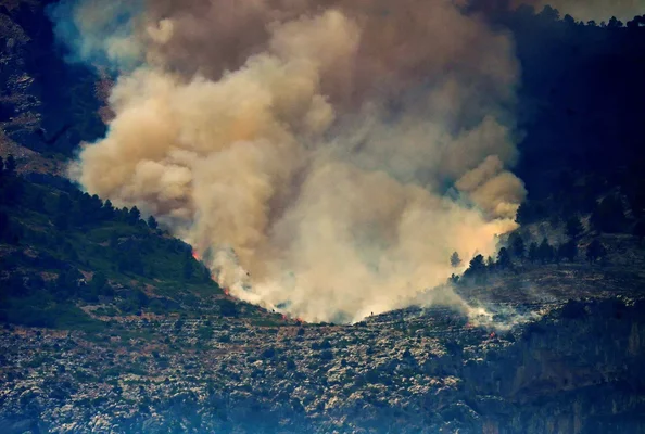 Un épais panache de fumée s'élève d'un incendie de forêt sur une montagne escarpée, avec des flammes visibles à la base.