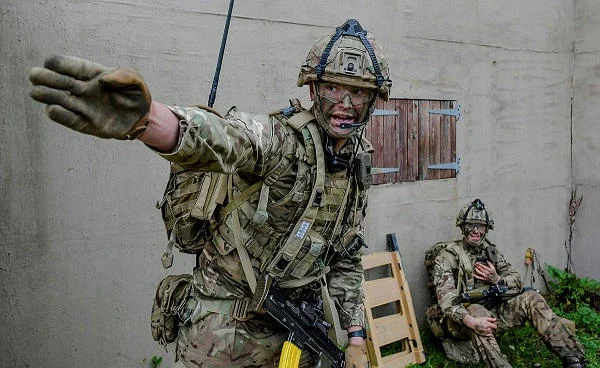 Soldat britannique en tenue de combat, faisant un geste de commandement, avec un autre soldat assis en arrière-plan.