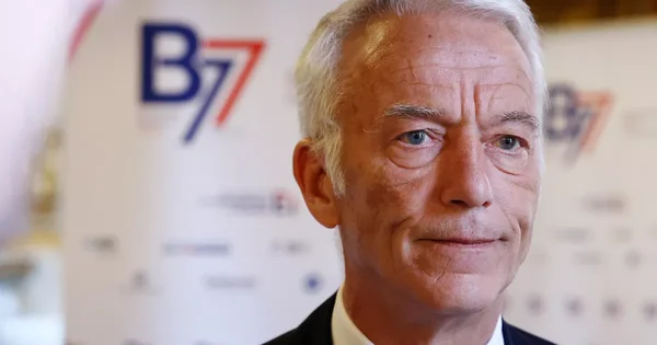Portrait d'un homme âgé devant un fond flou arborant le logo 'B77'.