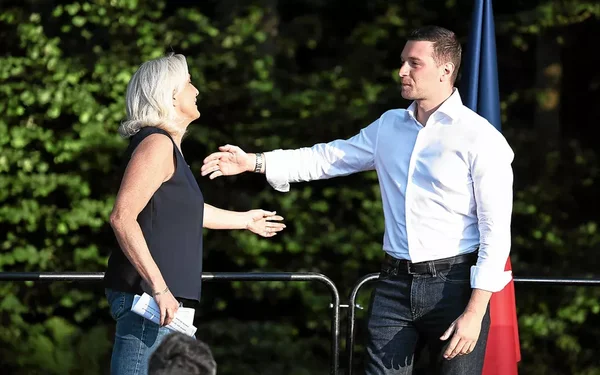 Jordan Bardella et Marine Le Pen se serrant la main en extérieur, devant un drapeau français.
