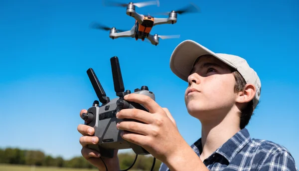 Gros plan sur les mains d'un adolescent tenant une manette de drone, regard concentré vers le ciel, drone quadricoptère visible en vol dans un champ dégagé, herbe et ciel bleu en arrière-plan, intention de montrer l'entraînement au pilotage de drone comme nouvelle compétence scolaire, ton sérieux et moderne