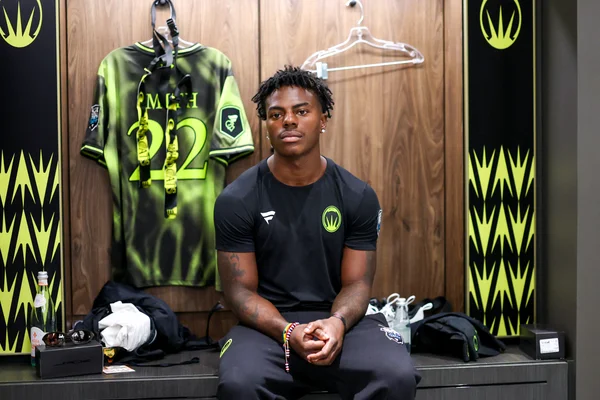 IShowSpeed dans un vestiaire, vêtu d'une tenue d'entraînement noire, un maillot vert et noir suspendu au mur.