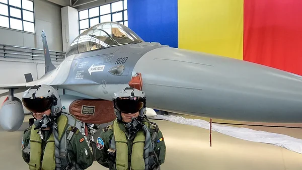 Deux pilotes roumains devant un F-16 dans un hangar, avec le drapeau national en arrière-plan.