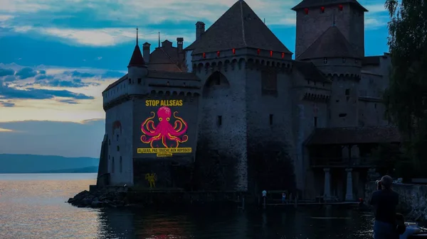 Projection d'une pieuvre rose et du message 'STOP ALLSEAS' sur le château de Chillon au crépuscule.