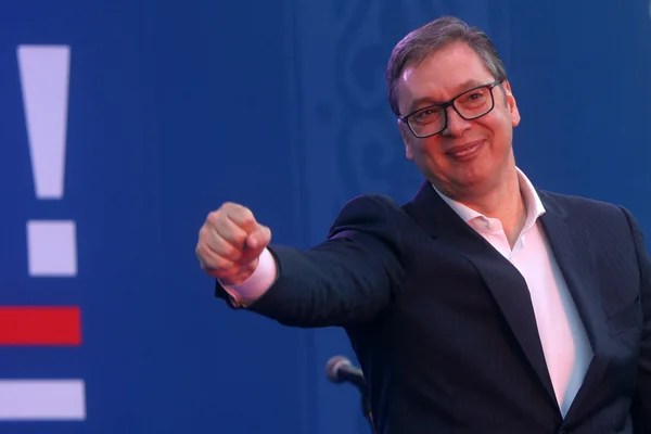 Le président Aleksandar Vučić faisant un geste lors d'une déclaration, souriant devant un fond bleu avec des éléments graphiques rouges et blancs.