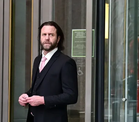 Carl Rinsch en costume noir et cravate rose ajustant sa veste devant une porte vitrée