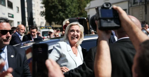 Marine Le Pen souriante, entourée de ses gardes du corps et de photographes dans un cadre urbain.