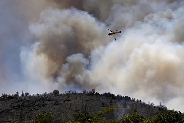 Un hélicoptère jaune avec un seau d'eau survole un incendie de forêt, laissant des terres brûlées au premier plan.