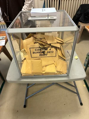 Urne transparente étiquetée 'CANTON 4 BUREAU 0413 RIGHI' remplie d'enveloppes jaunes dans un bureau de vote.