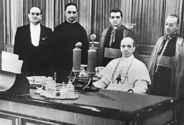 Le pape Pie XII lors d’une diffusion radiophonique, entouré de dignitaires ecclésiastiques.