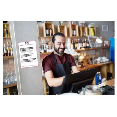 Un barman souriant derrière son comptoir, avec un panneau réglementaire « Vente d'alcool interdit aux mineurs » visible à l'arrière-plan.