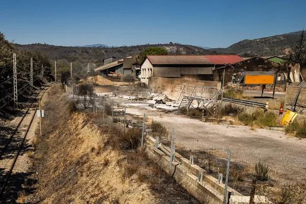 Site industriel adjacent à la voie ferrée, avec hangars et végétation sèche, après l'extinction des feux.