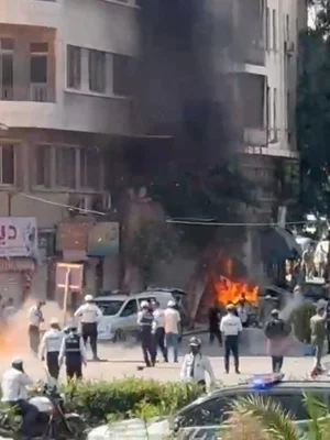 Véhicule en feu après une explosion à Damas, avec des policiers et des badauds sur les lieux.