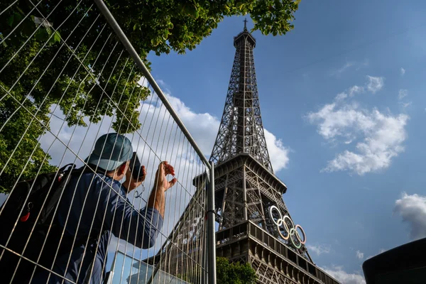Un touriste prend en photo la tour Eiffel avec les anneaux olympiques, derrière une grille de sécurité