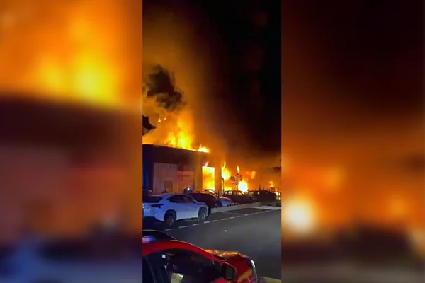 Incendie spectaculaire d'un garage automobile transformé en brasier géant, avec des voitures garées au premier plan.