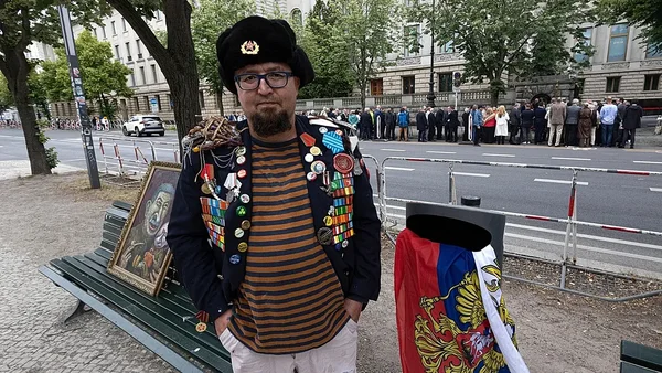 Semyon Skrepetsky, artiste russe et critique de Poutine, posant en tenue militaire ornée de médailles devant un portrait et un drapeau russe, entouré d'une foule.