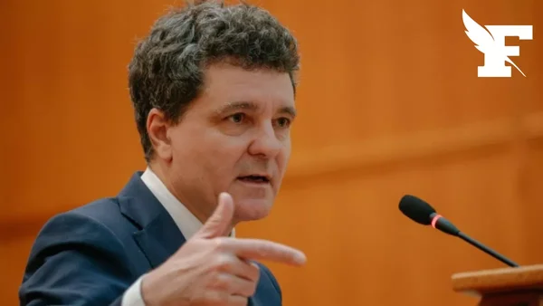 Un responsable s'exprime lors d'une conférence de presse, arrière-plan orange.