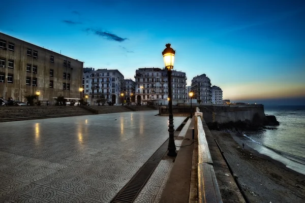 La promenade côtière d'Alger au crépuscule, avec la Casbah en arrière-plan.