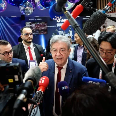 Jean-Luc Mélenchon entouré de micros lors d'un événement médiatique, faisant un signe de la main.