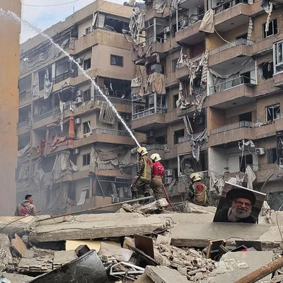 Pompiers intervenant sur un immeuble endommagé au Liban, avec un portrait de l'ayatollah Khamenei parmi les décombres.