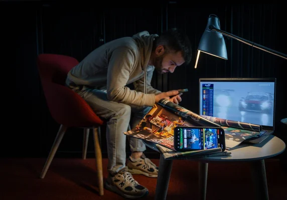 Homme en sweat beige examinant un poster, avec un ordinateur et un smartphone sur une table éclairée par une lampe.